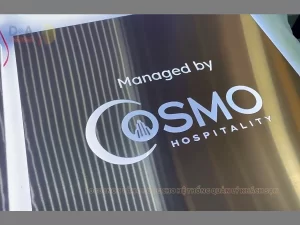 Logo inox vàng hồng xước COSMO Hospitality với nhiều chi tiết nhỏ, gia công tỉ mỉ trên nền bảng trang trí thương hiệu.