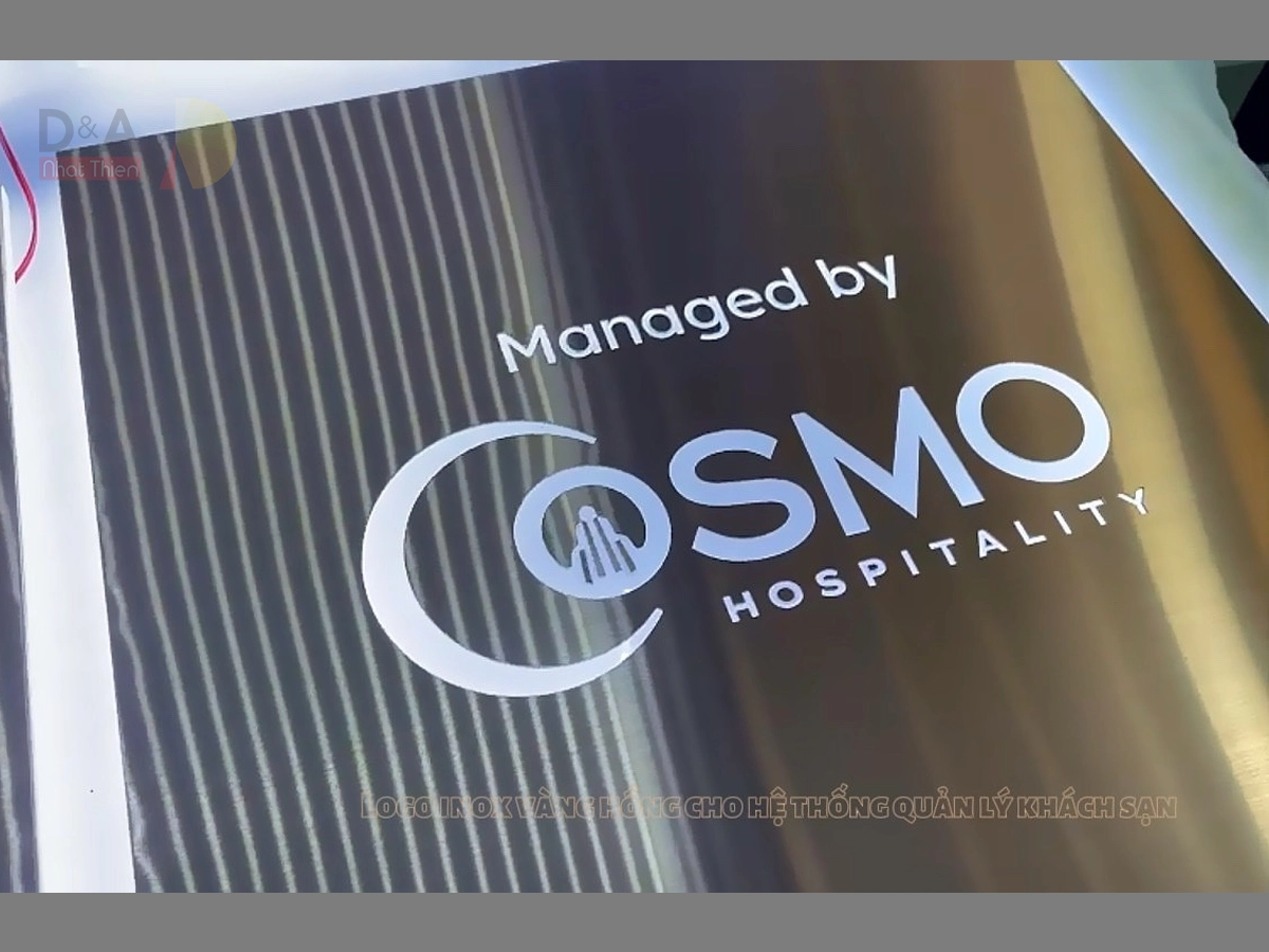 Logo inox vàng hồng xước COSMO Hospitality với nhiều chi tiết nhỏ, gia công tỉ mỉ trên nền bảng trang trí thương hiệu.